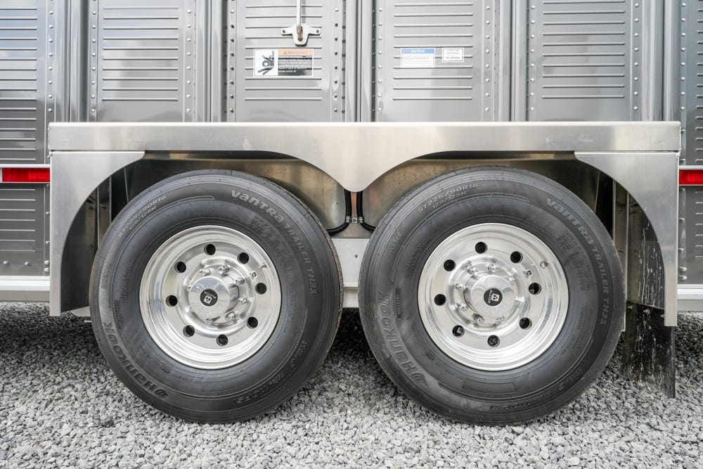 Twin tandem trailer wheels on a silver utility trailer over a gravel surface, showing metal fenders and hub caps.