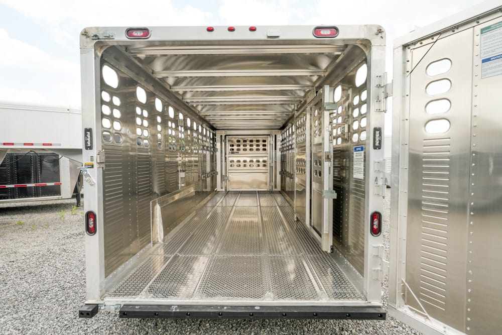 Interior of a polished metal livestock trailer, empty, with perforated side walls and grated floor lit by daylight from the rear doors