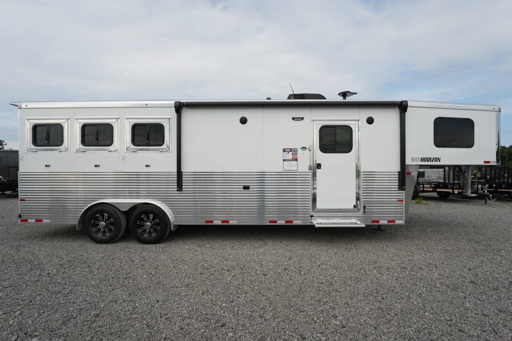 Silver aluminum horse trailer with three side windows, two axles, and a raised living-quarters section on the right, parked on a gravel lot.