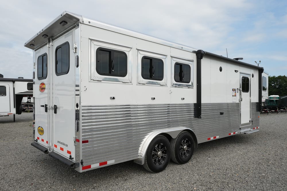 Enclosed white and silver horse trailer with three side windows and rear doors, parked on a gravel lot.