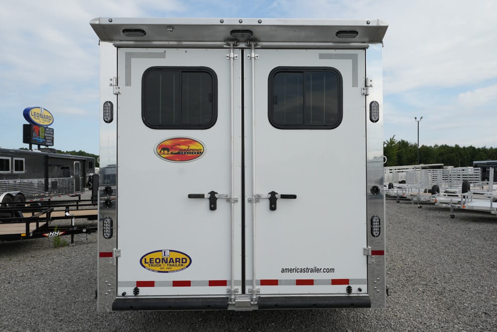 Rear view of a white cargo trailer with two doors, small windows, and brand stickers (Sundowner, Leonard) in a lot with other trailers nearby.