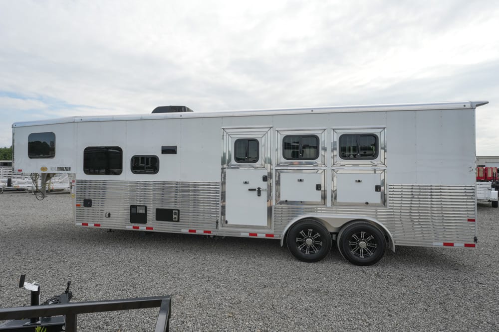 Silver aluminum horse trailer with two axles parked on a gravel lot; features multiple windows and side storage compartments as well as ribbed lower siding.