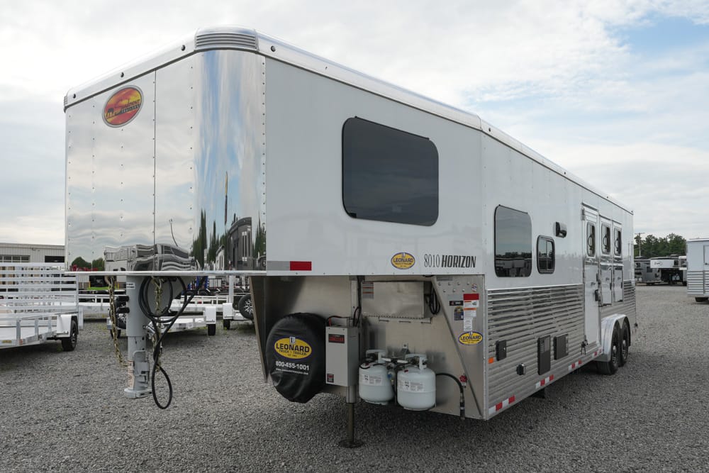Large silver fifth-wheel travel trailer on a gravel lot, front hitch extended and propane tanks visible.