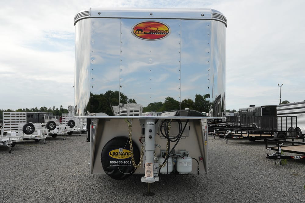 Shiny aluminum gooseneck horse trailer front with a spare tire and propane tanks visible beneath, parked in a gravel lot with other trailers nearby.