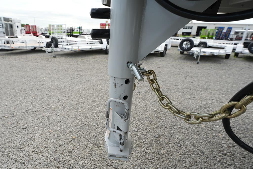 Close-up of a trailer tongue with a safety chain attached to the hitch on a gravel lot; other trailers in the background.