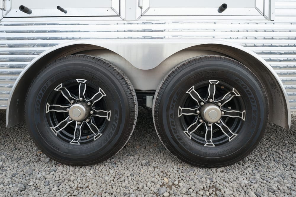 Pair of black trailer tires with stylish chrome-spoked rims under a shiny metal trailer body on gravel ground