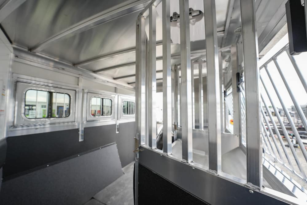 Interior of a metal livestock trailer with vertical dividers and windows along the side, sunlight filtering in from outside.