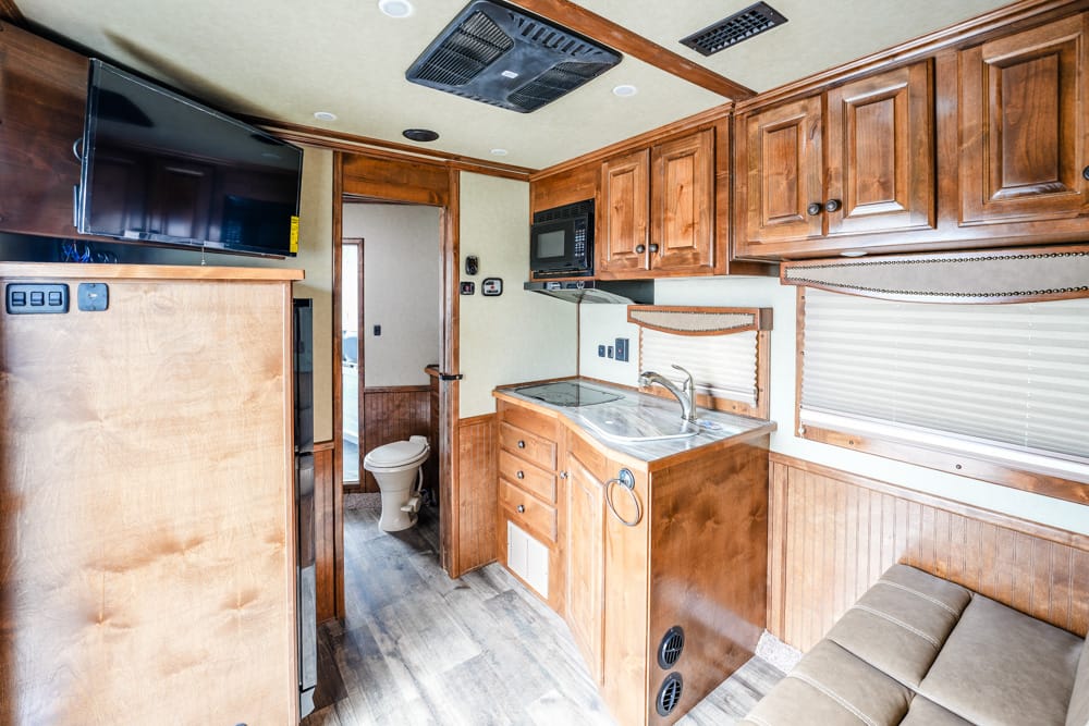 Interior of a wood-paneled RV kitchen with sink, stove, microwave, and cabinets; TV mounted above, sofa to the right, and a bathroom door visible in the back.