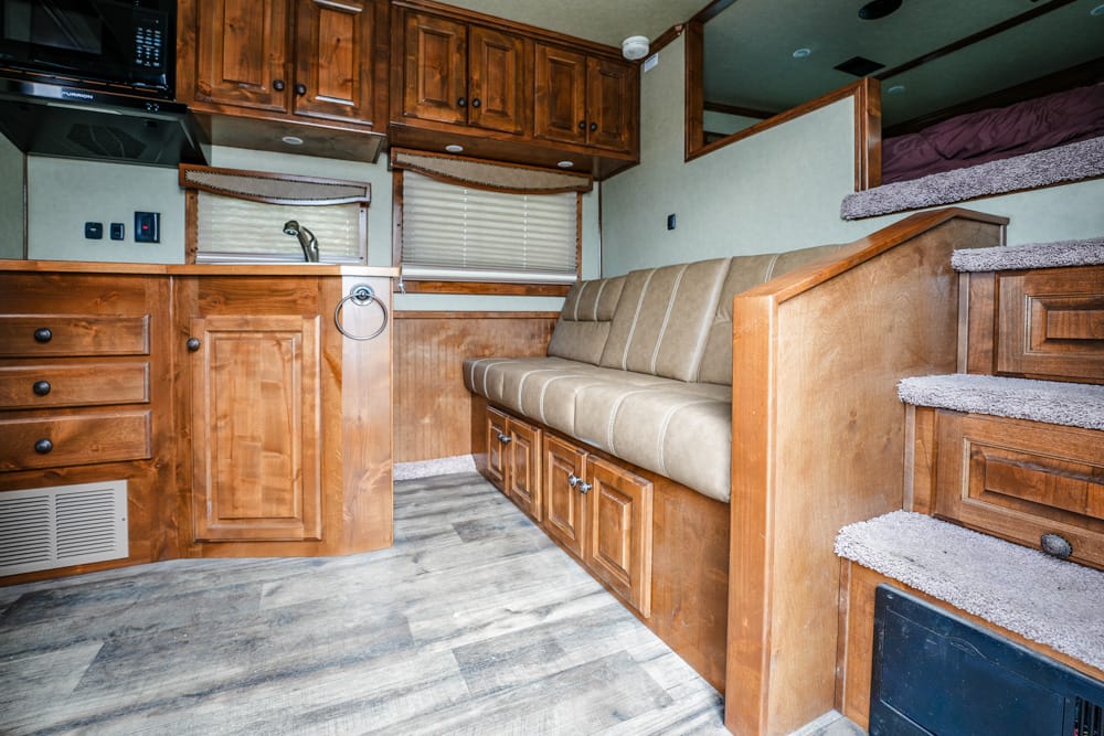 Wood-paneled RV interior with a beige cushioned sofa, wooden cabinets, and stairs leading up to a loft area.
