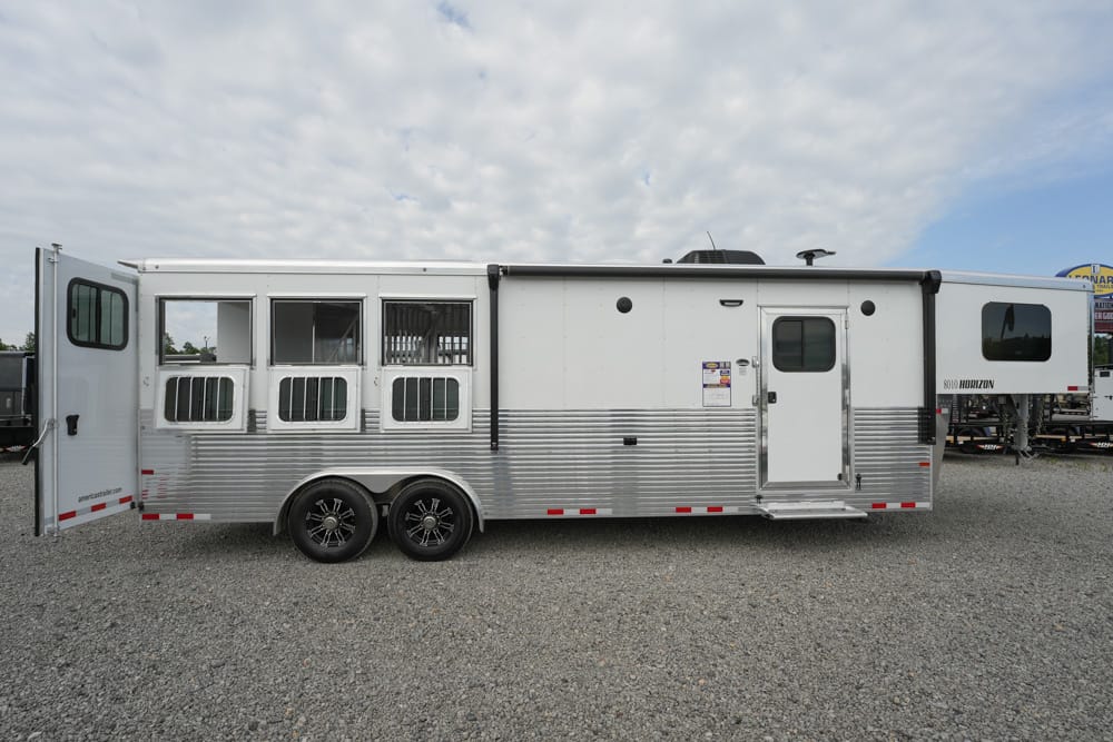 Silver aluminum horse trailer with two axles and multiple vented windows parked on a gravel lot.
