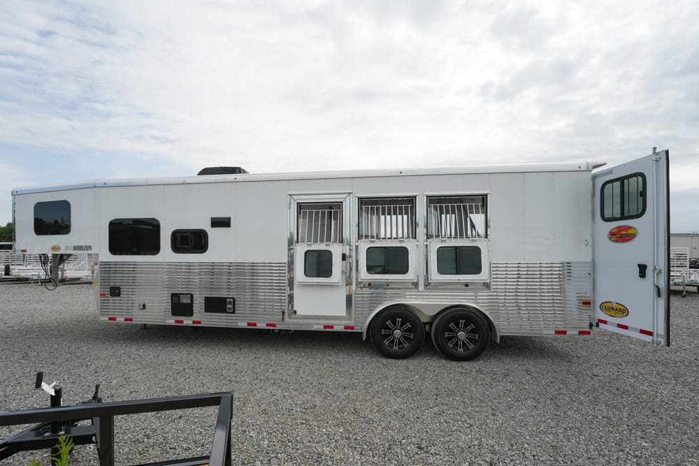 White aluminum horse trailer with three stalls, side windows, and a rear door open, parked on a gravel lot.