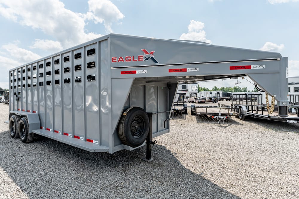 Gooseneck livestock trailer: long gray Eagle X trailer with ventilation slats and a spare tire mounted at the front, parked on gravel.