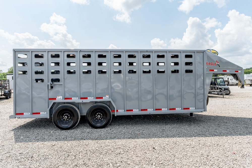 Grey livestock trailer with ventilation slats parked on a gravel lot, two-axle wheels visible, Eagle brand on the front, under bright sunny skies with fluffy clouds.