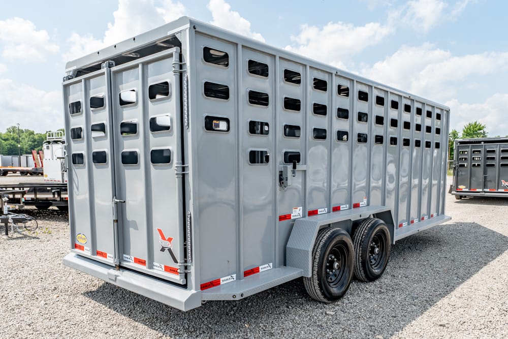 Gray livestock trailer with dual axles parked on gravel, back doors closed.Consise and informative for conveying purpose and setting.]