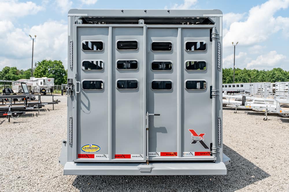 Rear of a gray livestock trailer with vented doors and brand decals parked on a gravel lot.