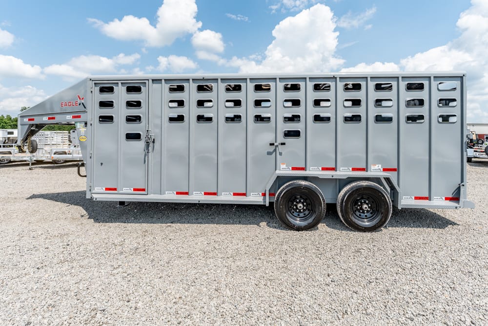 Gray livestock trailer with vented side panels and dual rear wheels on a gravel lot, under a blue sky with clouds.