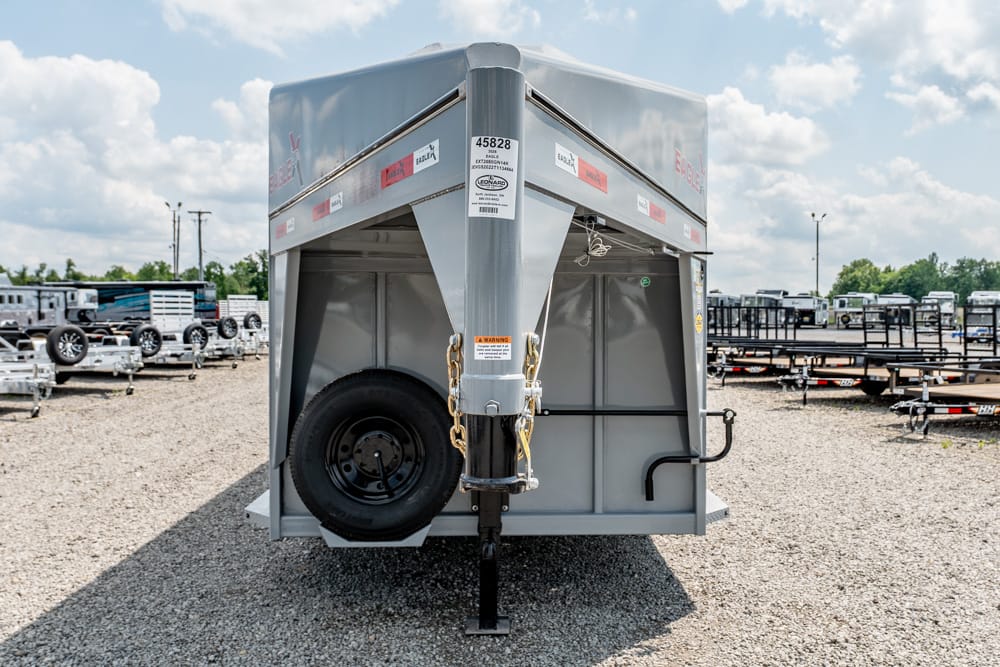 Front view of a large silver utility trailer with a spare tire mounted on the left, safety chains, and a hitch, parked on gravel in a trailer lot.