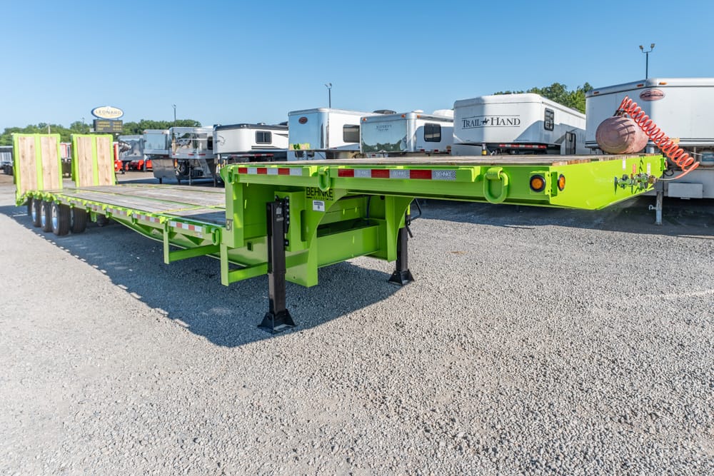 Lime-green heavy-duty flatbed trailer on deployed support legs in a gravel lot with other trailers in the background.