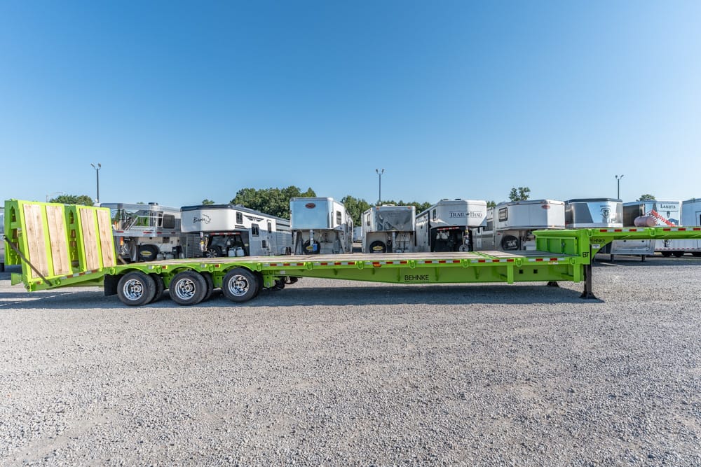 Green lowboy trailer with multiple axles parked on a gravel lot beside row of travel trailers/RVs in the background.