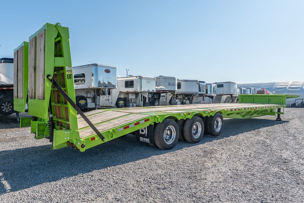 Green flatbed trailer with folding loading ramps on a gravel lot, carrying several white travel trailers in the background.