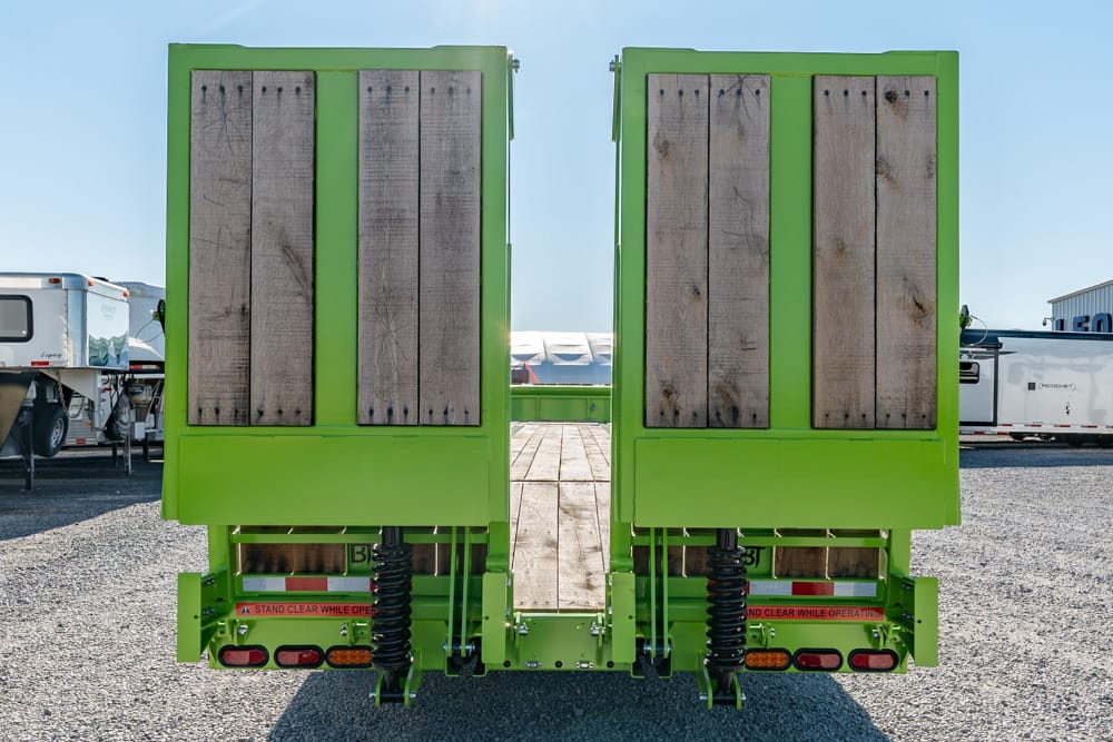 Rear view of two bright green trailers with wooden side doors, parked on a gravel lot with RVs in the background.