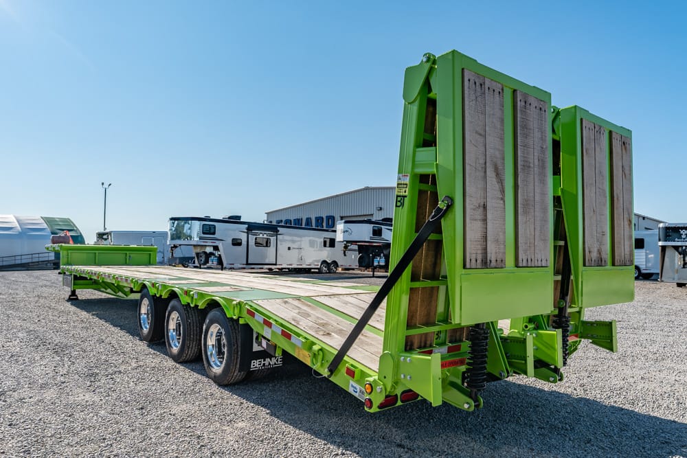 Green heavy-duty flatbed trailer with rear ramps in a gravel lot, RVs and buildings in the background.