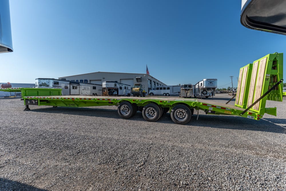 Bright green lowboy trailer with a rear ramp in a gravel yard, with trucks and a building in the background.