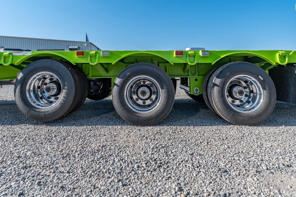 Green heavy-duty trailer chassis with three axles and chrome wheels on gravel under clear blue sky.