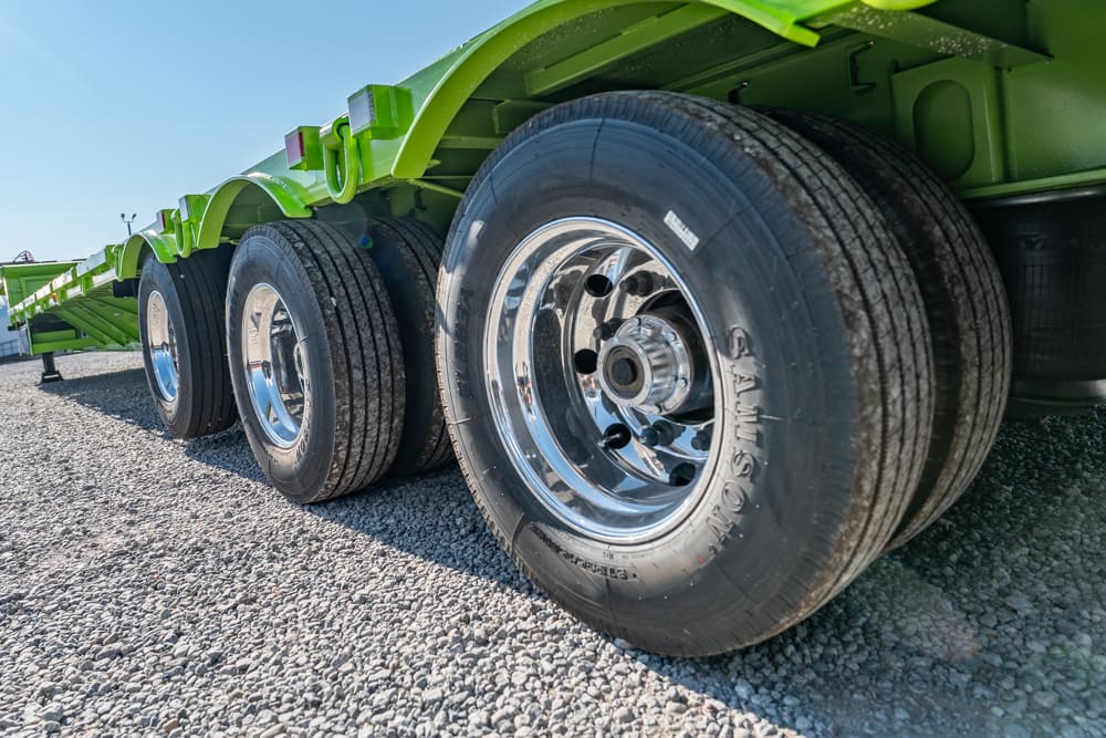 Close-up of steel wheels and tires on a green flatbed trailer parked on gravel surface at a low angle.