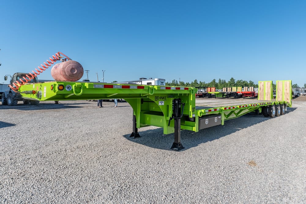 Bright green heavy-duty lowboy trailer on a gravel lot with a coiled orange air hose and wooden blocks at the far end.
