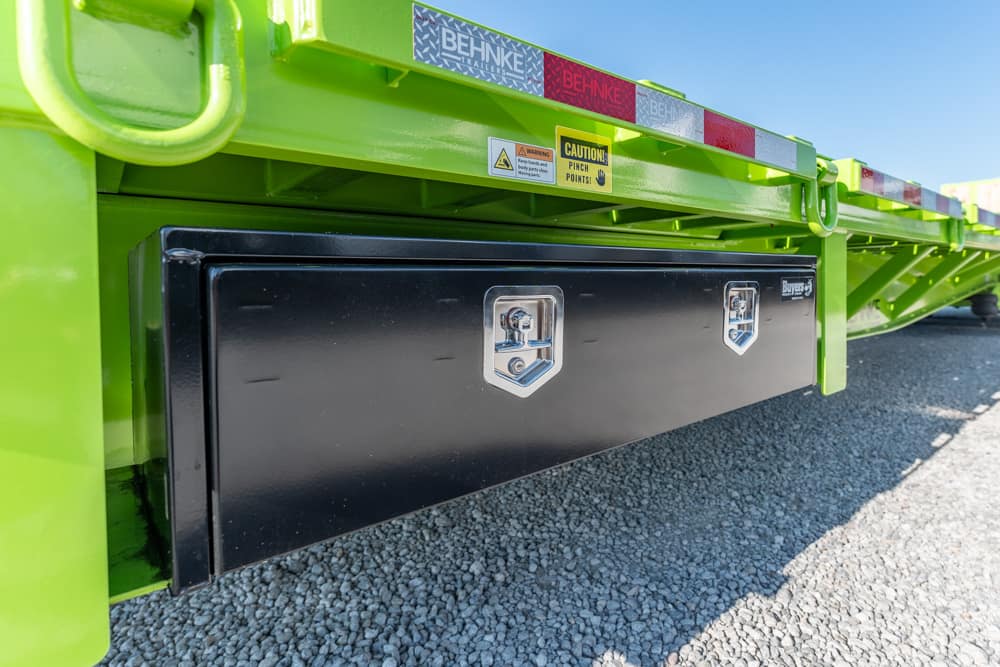 Bright green trailer with a large black undercarriage toolbox and metal latches, on gravel surface.