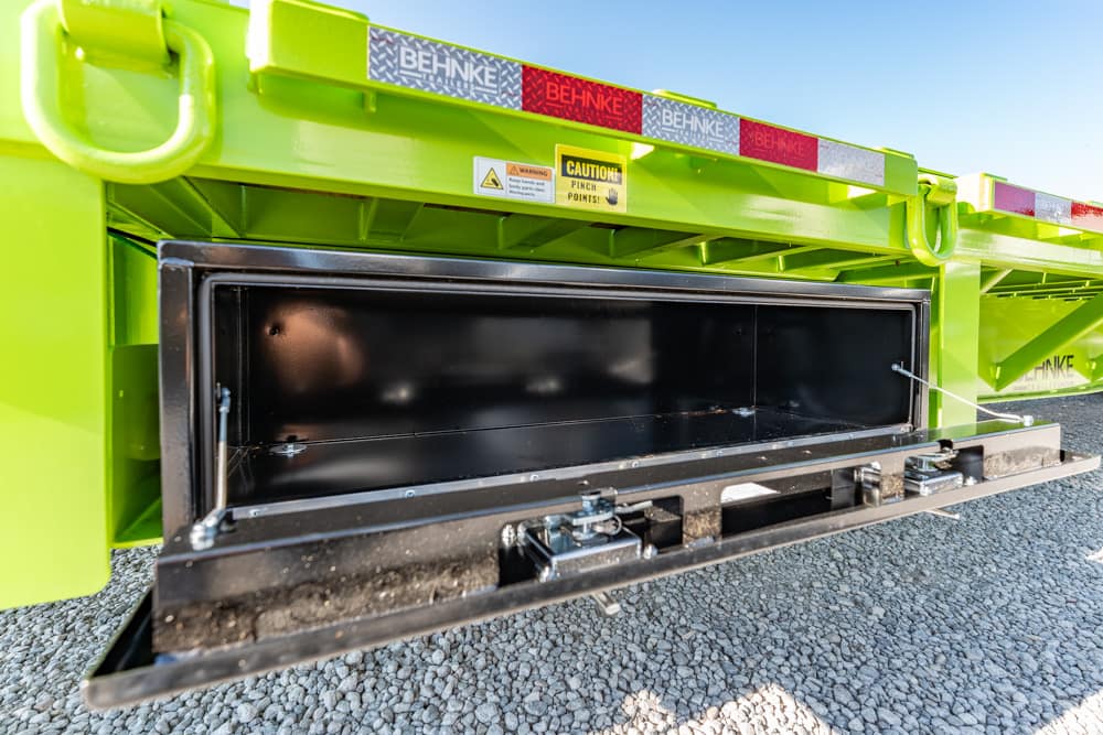 Open storage hatch on a bright green Behnke trailer, metal interior and latch mechanisms visible.
