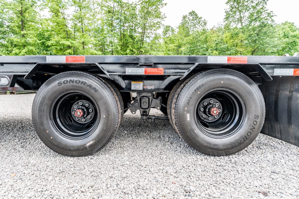 Close-up of a dual-axle trailer with black wheels and heavy-duty tires on gravel, greenery in the background.