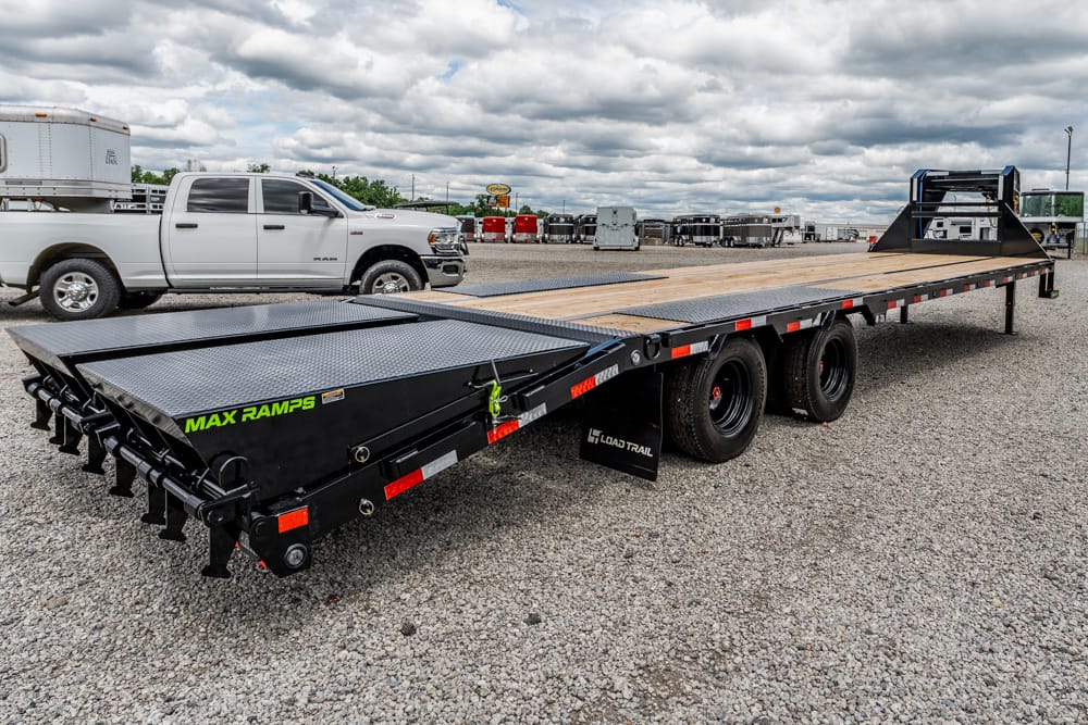 Wide shot of a black MAX RAMPS flatbed trailer with a diamond-plate deck and rear loading ramps, parked on a gravel lot beside a white pickup truck.