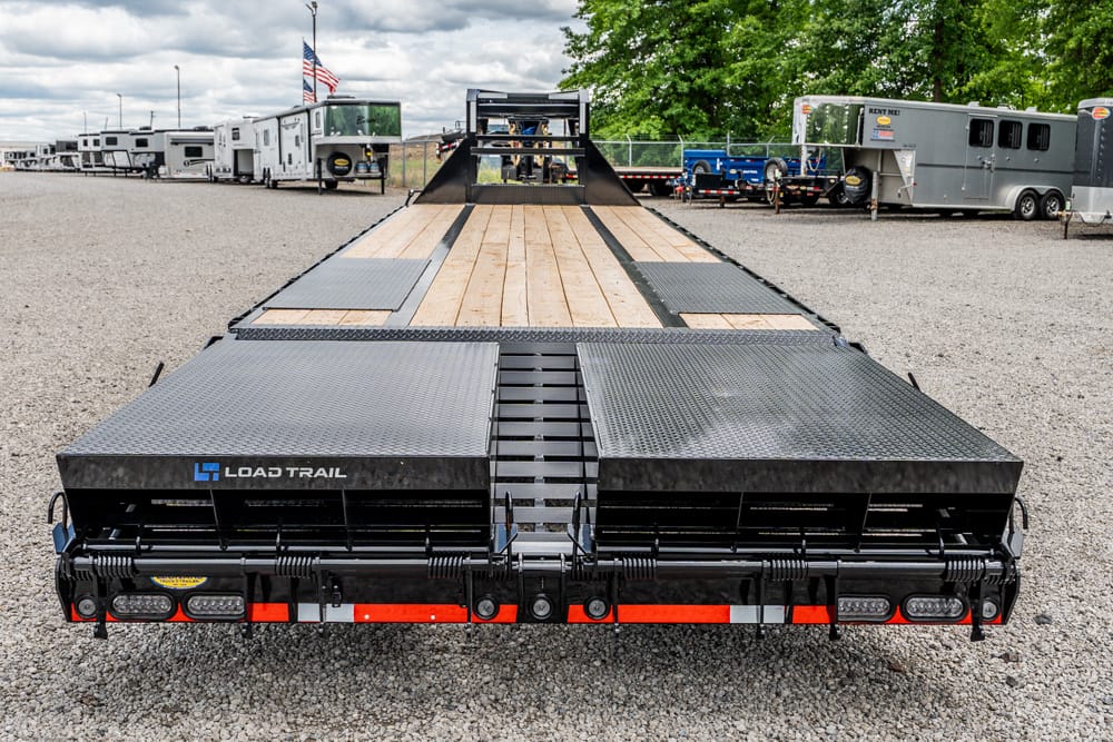 Long flatbed trailer with a wooden deck and metal ramps, parked on a gravel lot with other trailers in the background (Load Trail branding visible).