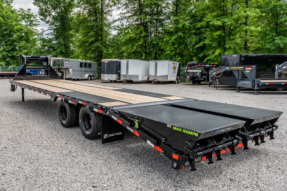 Flatbed trailer with dual ramps labeled Max Ramps in a gravel lot, trees in the background, ready for loading equipment.