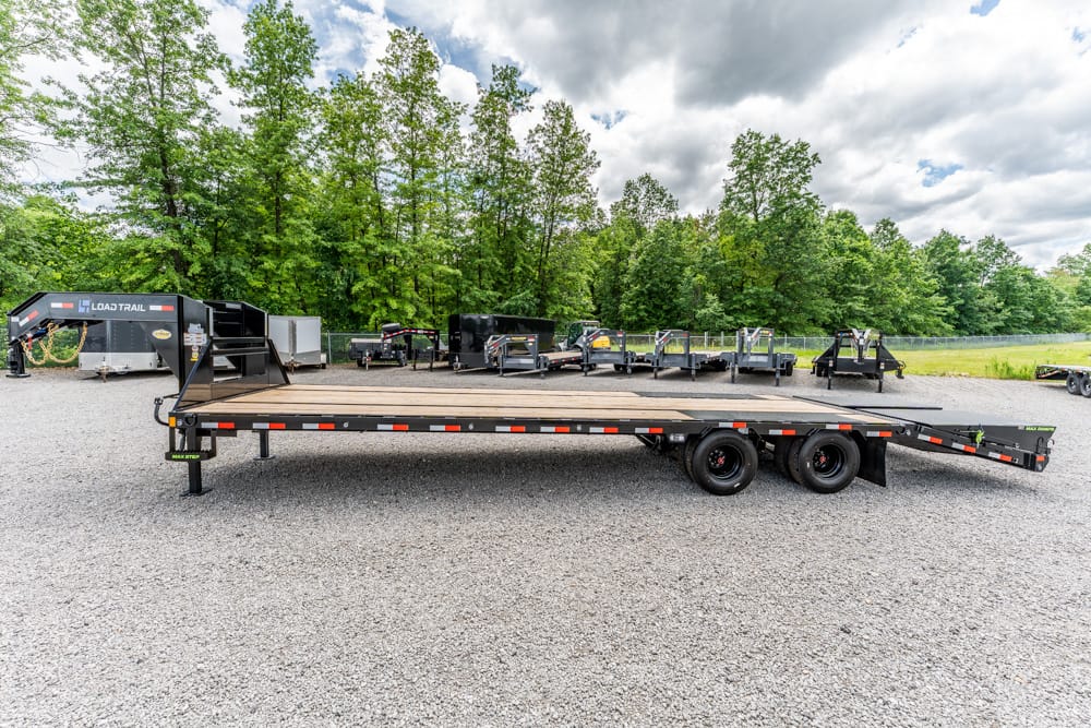 Long flatbed trailer on a gravel lot with trees and other equipment in the background.