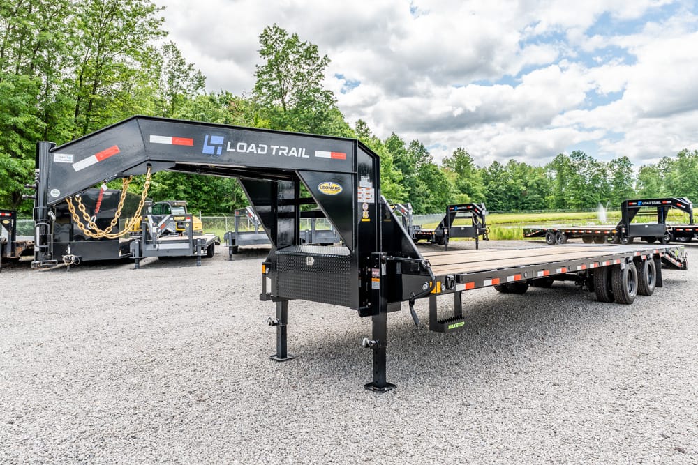 Gooseneck flatbed trailer on a gravel lot, branded Load Trail, with support legs deployed and trees in the background