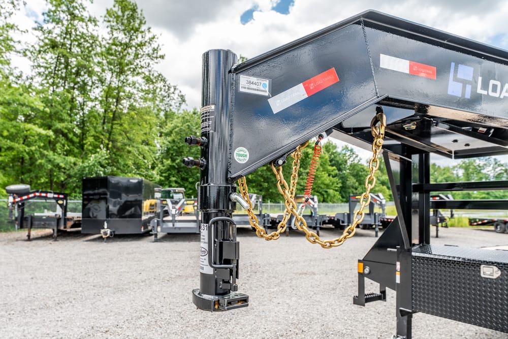 Gooseneck trailer hitch with yellow safety chains attached, in a gravel lot with construction trailers in the background.