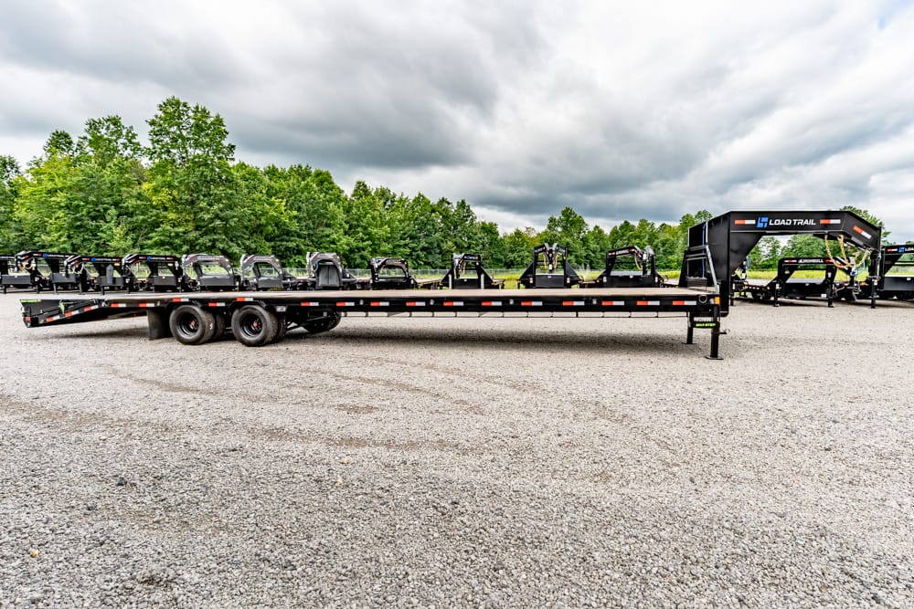 Long black flatbed trailer with tandem wheels and loading racks parked on a gravel lot, Load Trail logo visible against a cloudy sky and trees in the background.