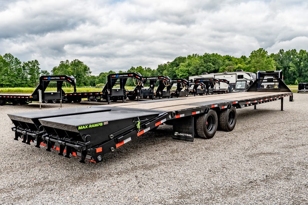 Long black Load Trail flatbed trailer with rear ramps in a gravel lot, with other trailers in the background.