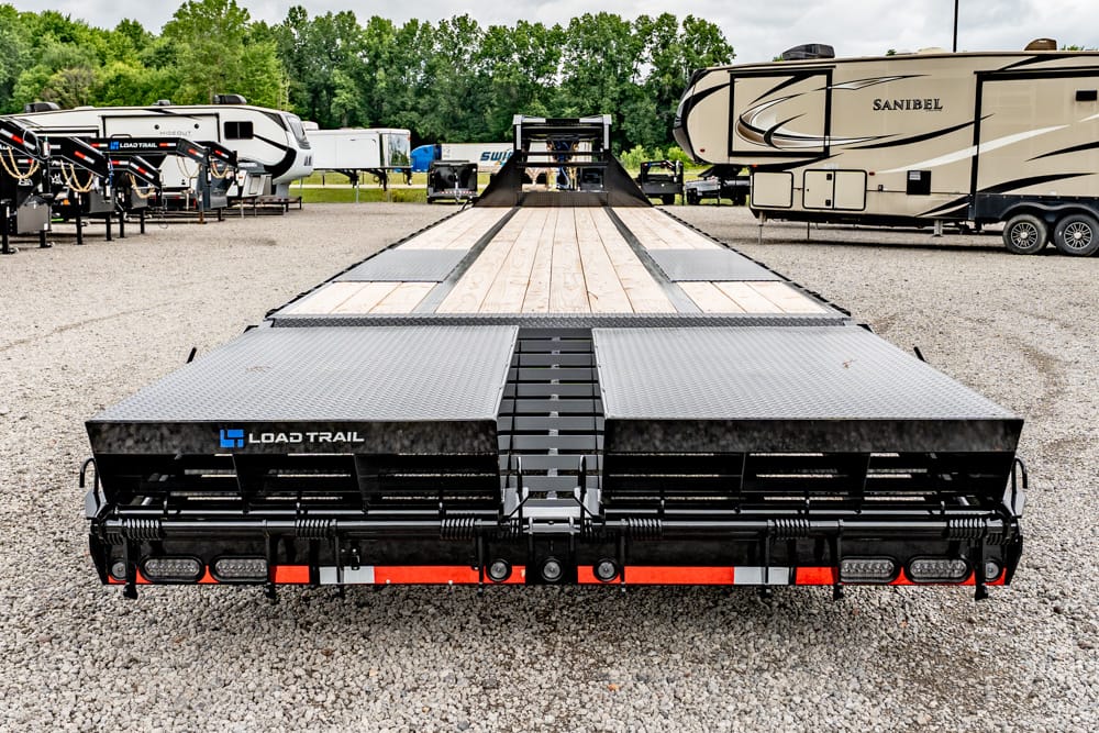 Rear view of a Load Trail flatbed trailer with a wooden deck and ramp sections, parked in a gravel RV lot with campers in the background.