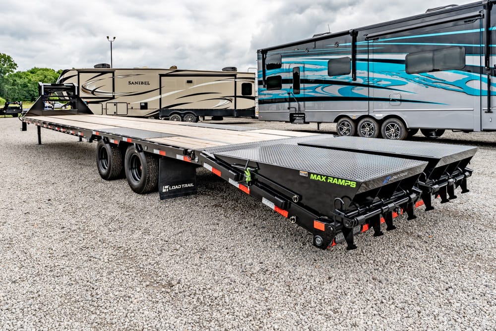 Long black Load Trail flatbed trailer with ramps, parked on a gravel lot beside large RVs.