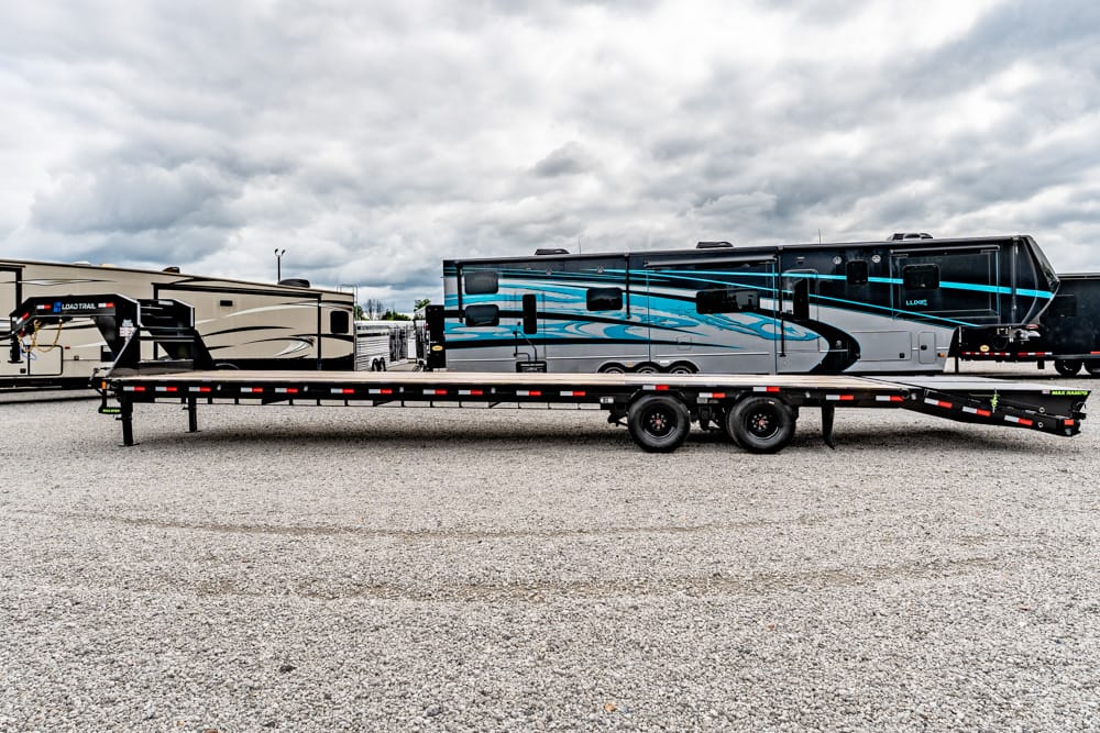 Blue-and-gray luxury fifth-wheel RV loaded on a long black flatbed trailer in a gravel lot under a cloudy sky.