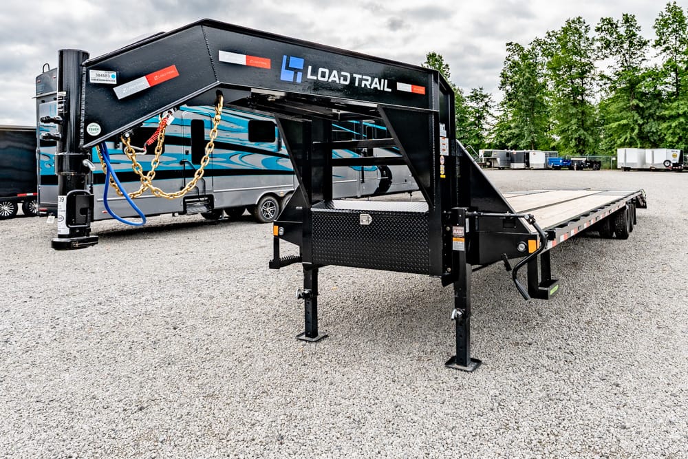 Black Load Trail gooseneck flatbed trailer on a gravel lot with trees in the background, ready for hauls