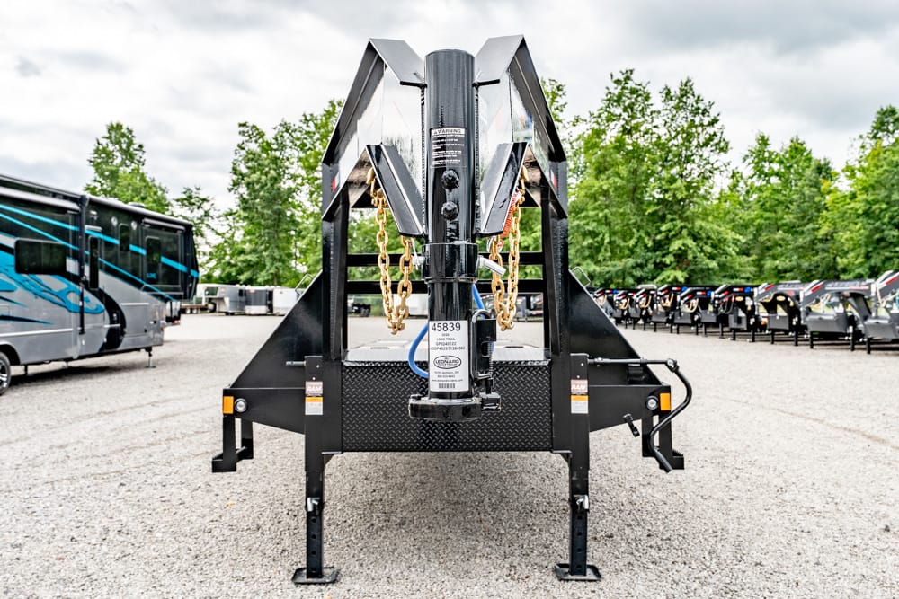 Front view of a heavy-duty black gooseneck trailer with safety chains and hitch, parked on gravel with RVs and trees in the background.