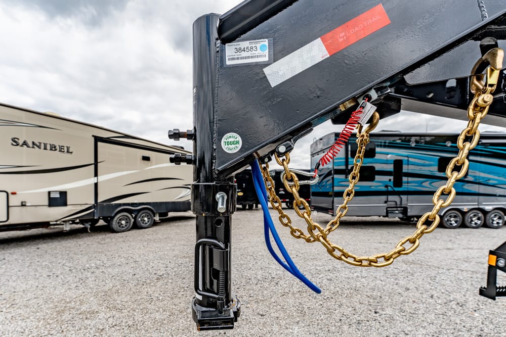 Close-up of a gooseneck trailer hitch with gold safety chains and blue electrical cables in a gravel lot of RVs behind it.