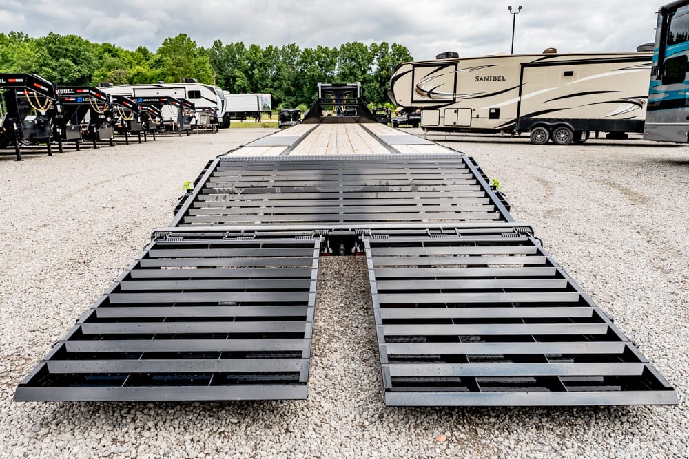 Long, low-angle view of a black metal car trailer ramp with wooden deck sections, parked on gravel with RVs in the background