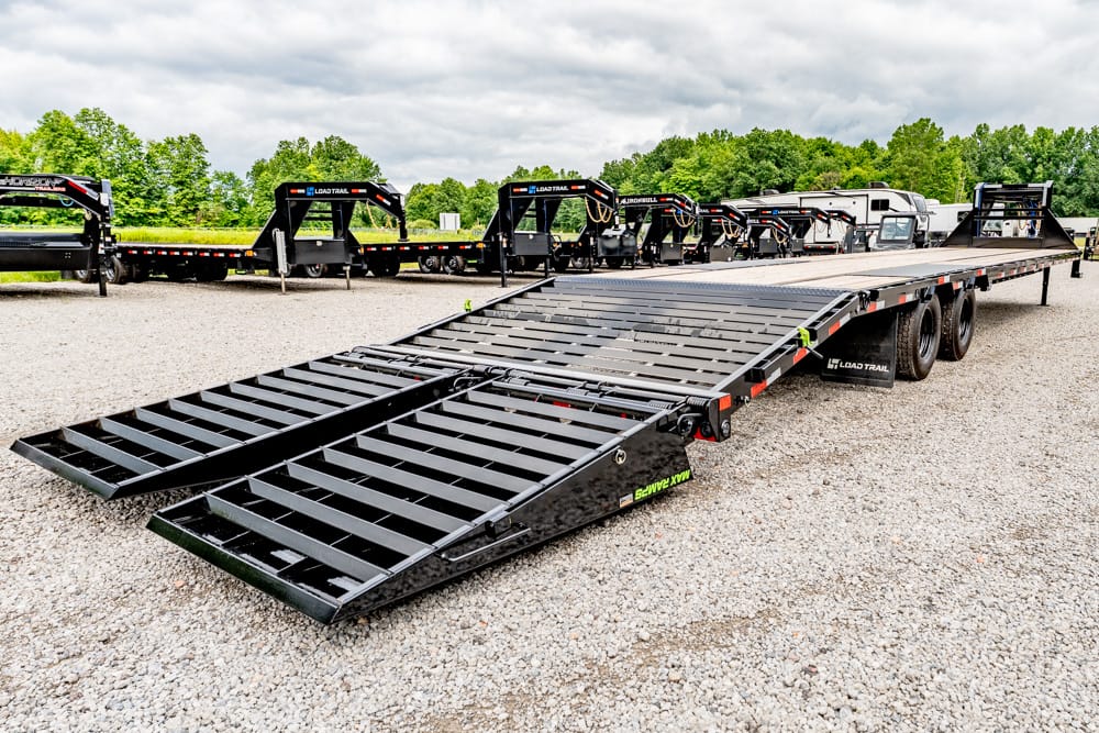 Black heavy-duty car trailer with two fold-down ramps on a gravel lot, others lined up in background under a cloudy sky