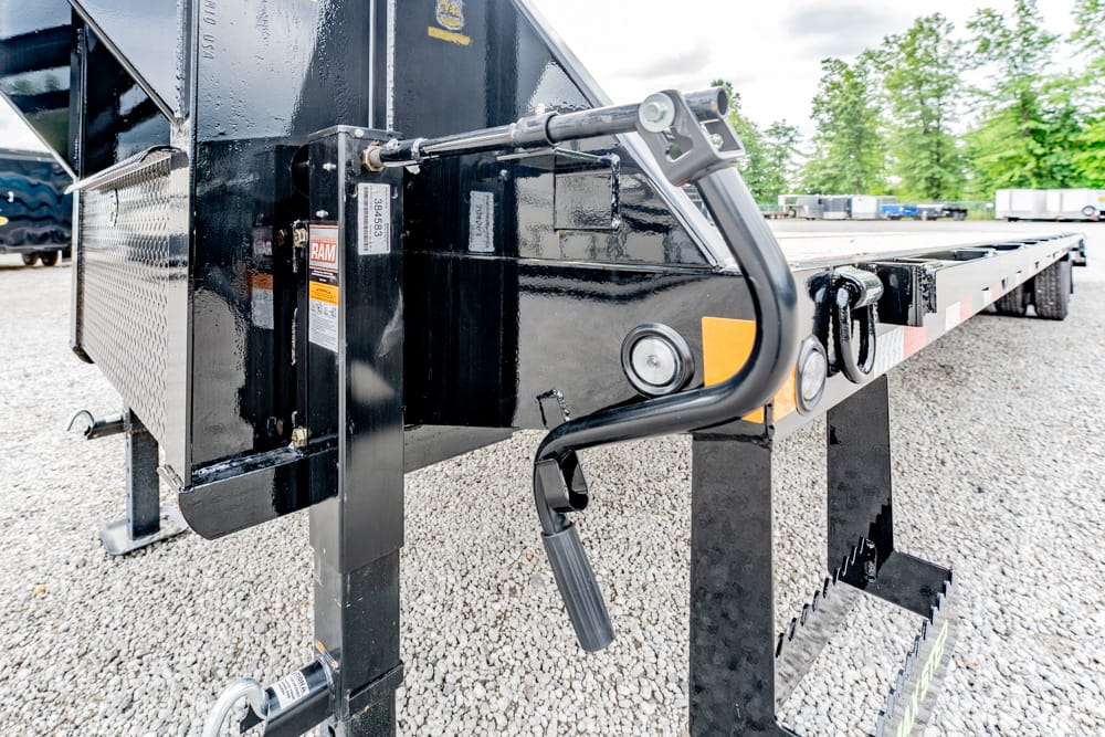 Close-up of a black flatbed trailer hitch with a crank jack and support leg on a gravel lot, with other trailers in the background